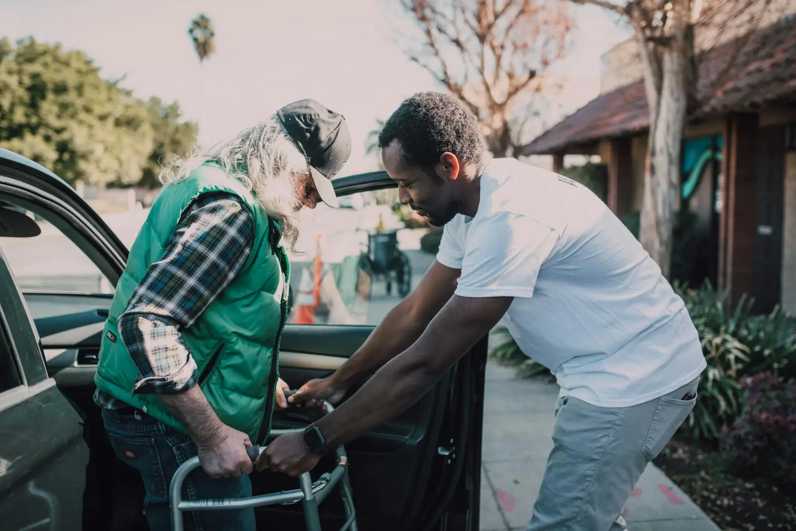 Older adult using a walker while a helper steadies the car door in a parking area